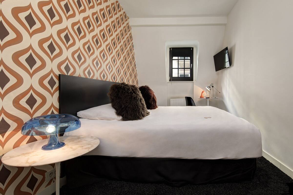 Ein modernes Hotelzimmer mit einer Wand mit Muster, einem Bett mit schwarzem Kopfteil, einem kleinen Nachttisch mit Lampe und einem an der Wand befestigten Fernseher.