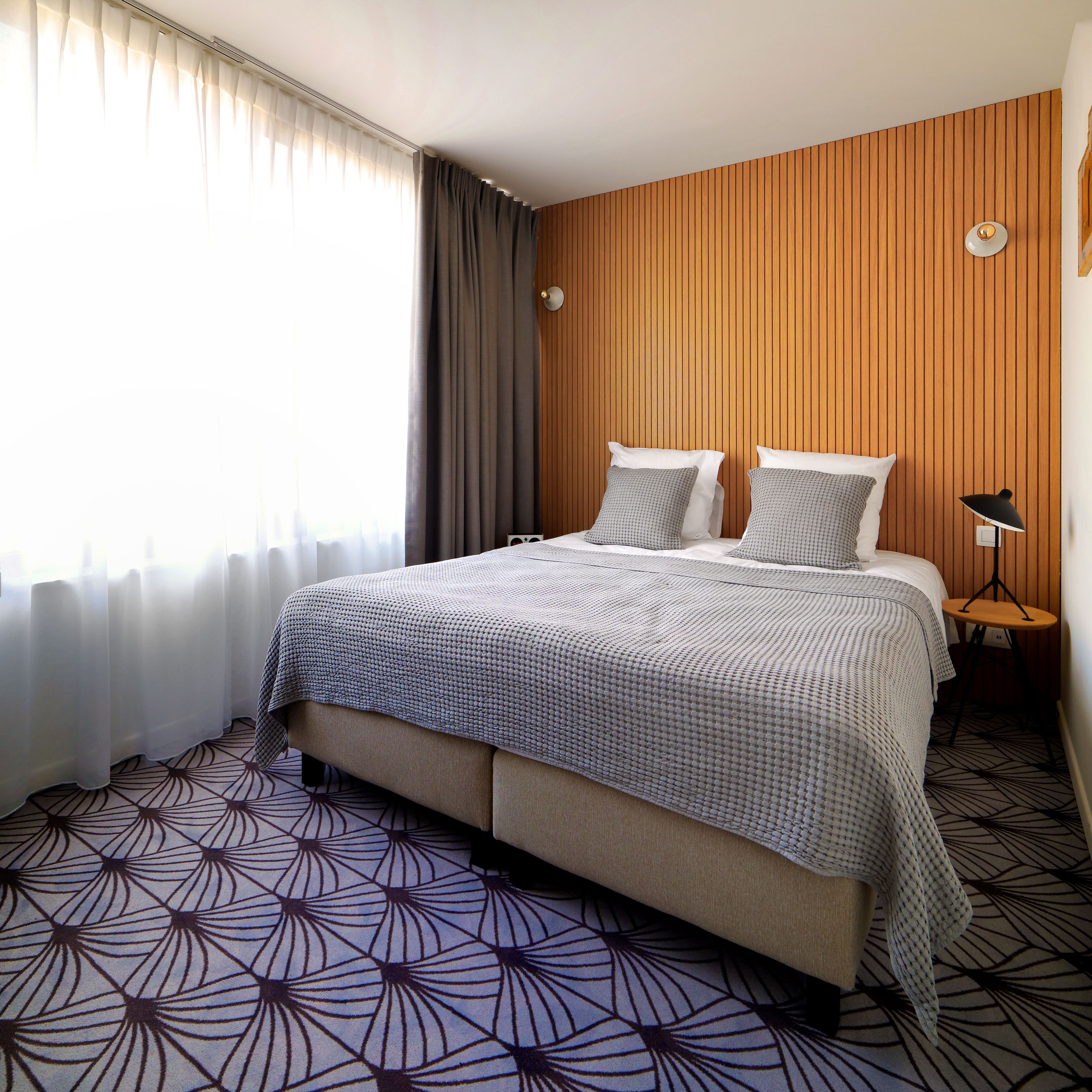 A hotel room with a bed, curtains, a wooden panel wall, a small table with a lamp, and a patterned floor.