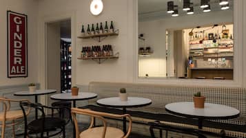 Een gezellig bargedeelte met ronde tafels, rieten stoelen en een bank. Boven de bar staan planken met verschillende flessen.