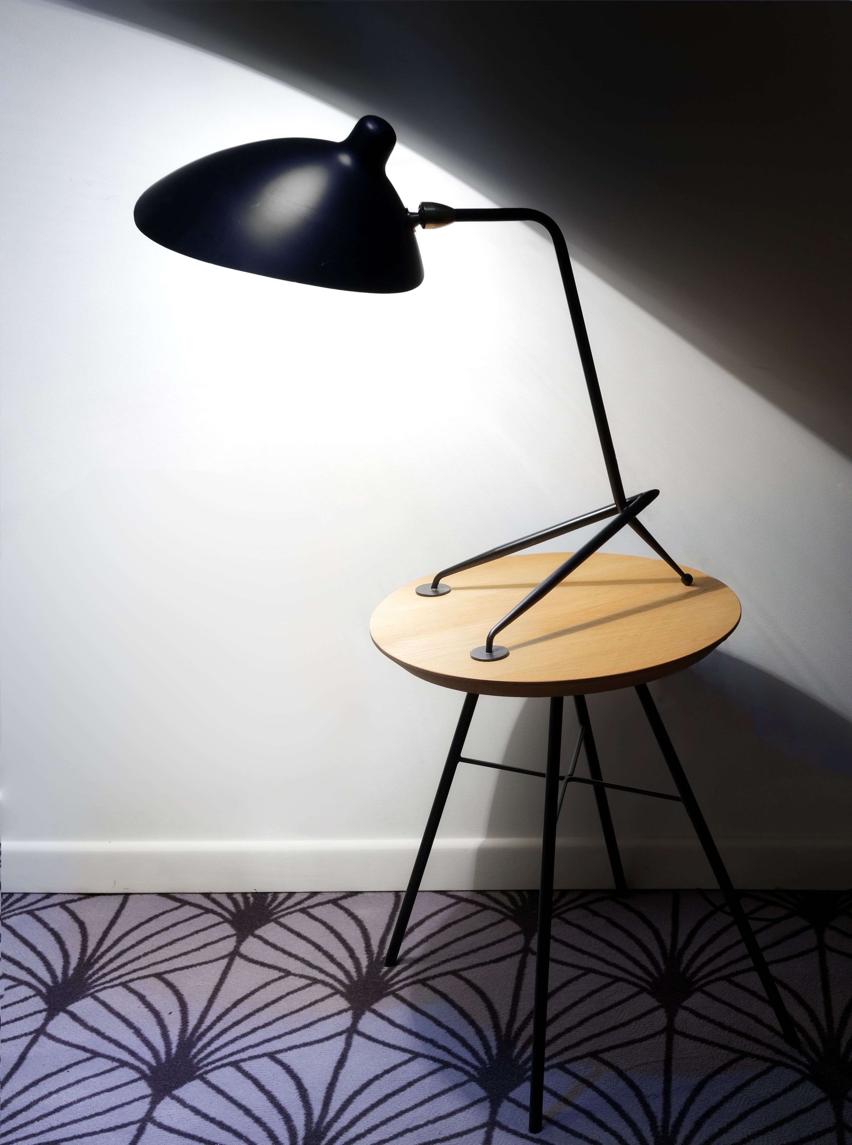 Een moderne stoel met een houten zitvlak en zwarte metalen poten, geplaatst voor een muur met een geometrisch vloerkleed.