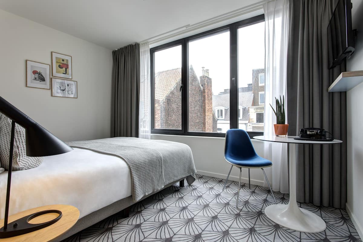 Ein modernes Hotelzimmer mit Bett, Schreibtisch, Stuhl und großem Fenster mit Blick auf die Stadt.