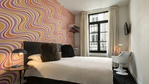 A hotel room with a large bed, a bedside table with a lamp, a chair, a window with curtains, and a colorful wall.