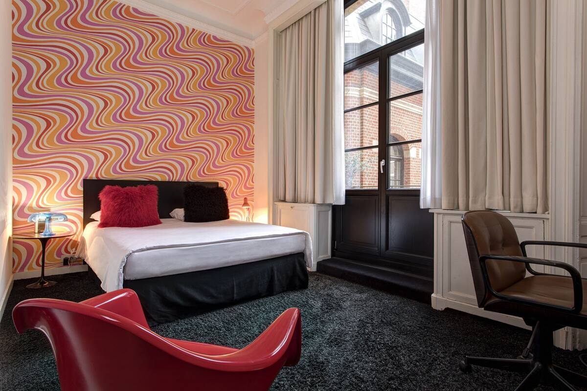 Ein Hotelzimmer mit einem Bett, einem roten Sessel, einem Schreibtisch, einem Fenster mit Vorhängen und einer Wand mit Muster.
