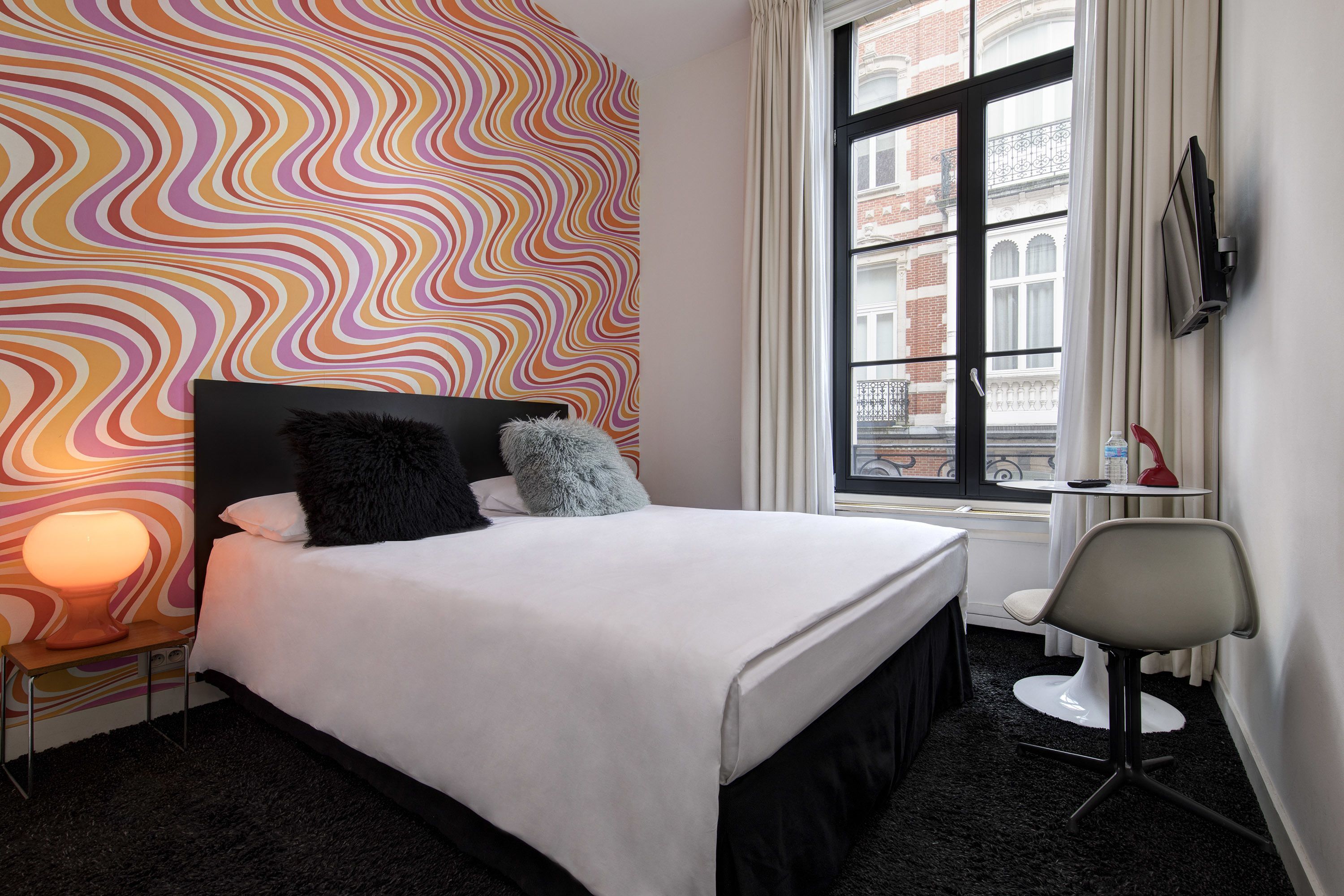 Habitación de hotel con cama, una silla, televisión y ventana con vista a edificios.