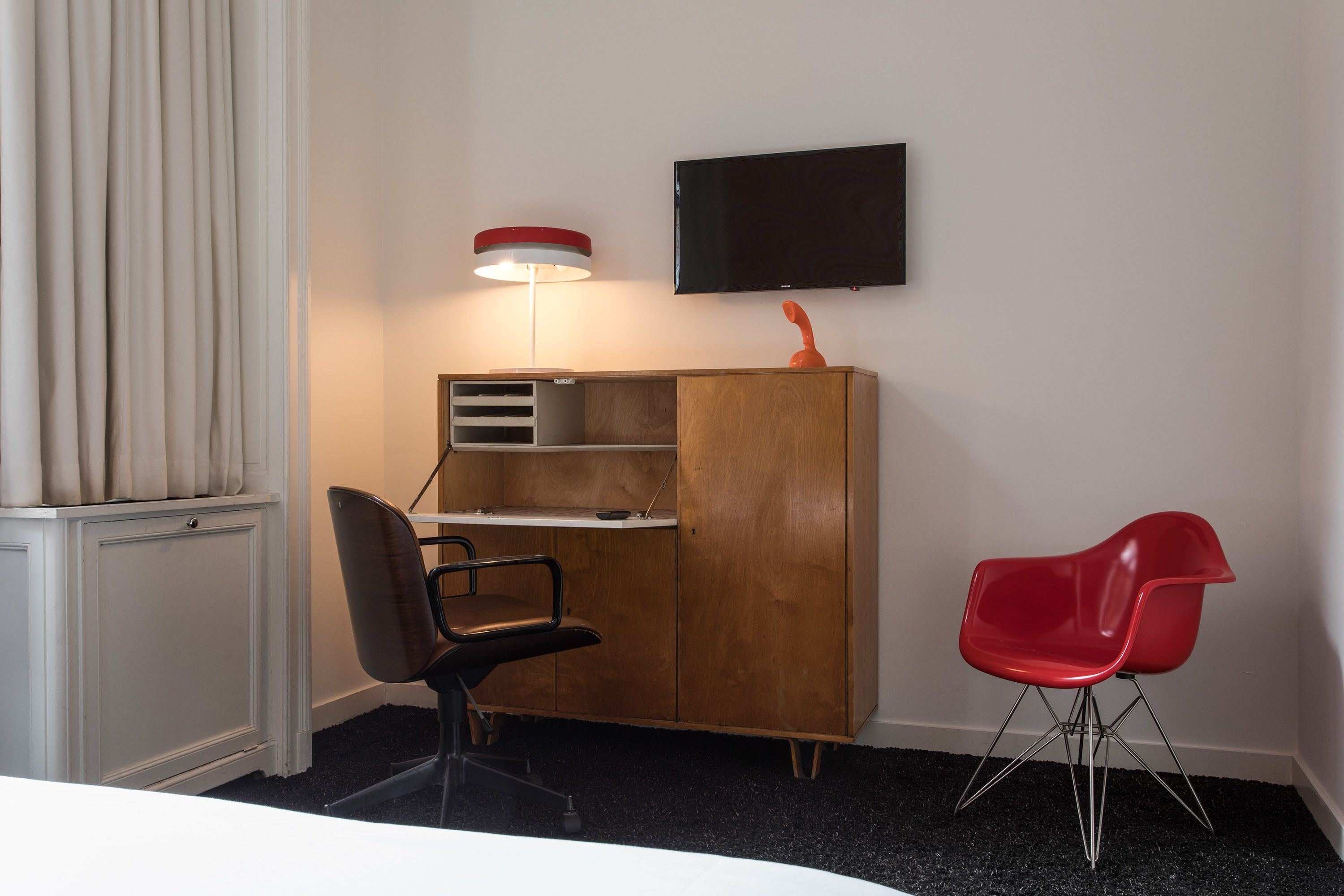 A hotel room with a wooden desk, a red chair, a television on the wall, and a window with curtains.