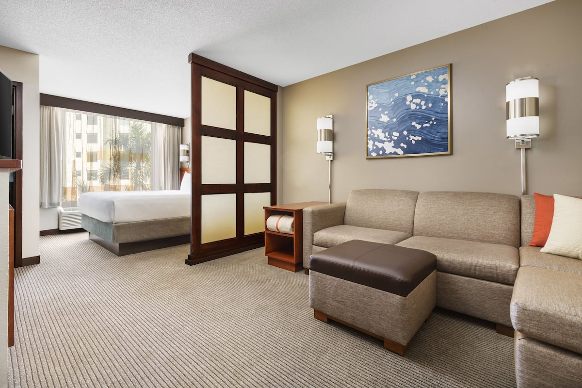 A hotel room with a bed, sofa, ottoman, and a large window with curtains.