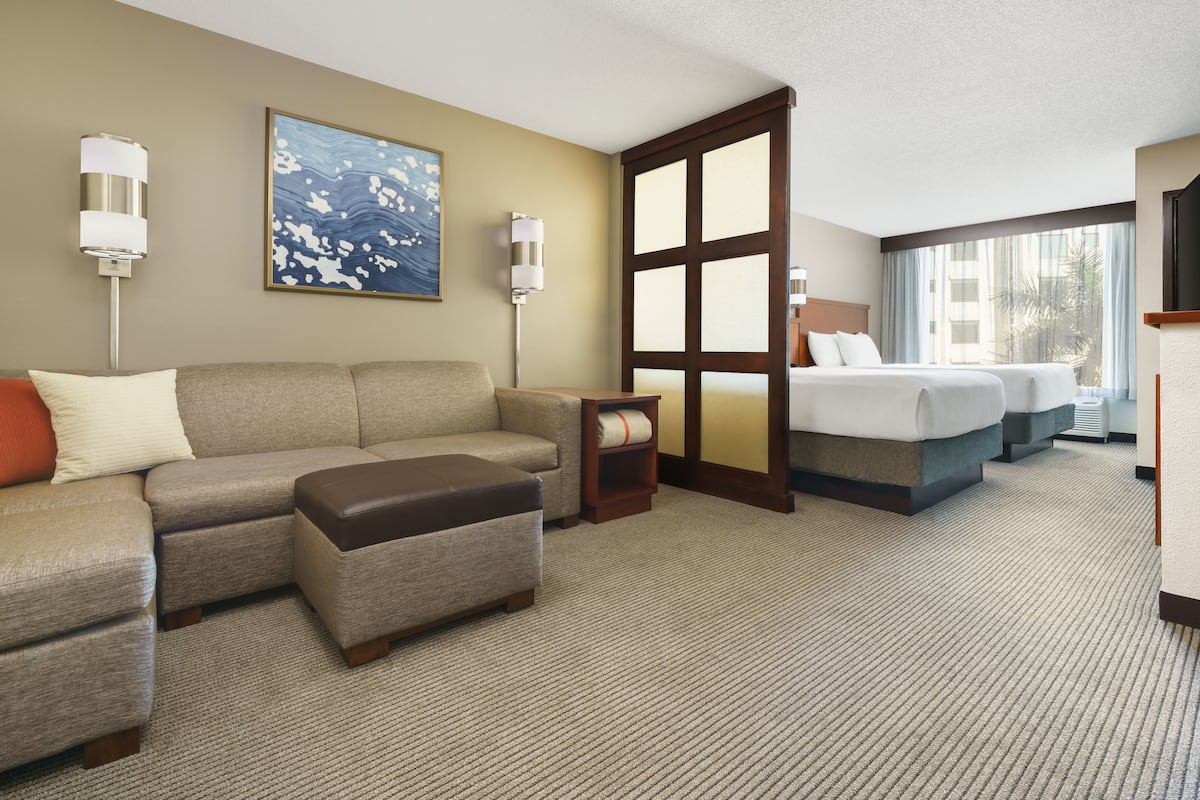 A hotel room with a large sectional sofa, a small ottoman, a bed with two pillows, a nightstand, and a large window with curtains.