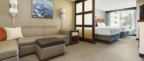 A hotel room with a large sectional sofa, a small ottoman, a bed with two pillows, a nightstand, and a large window with curtains.
