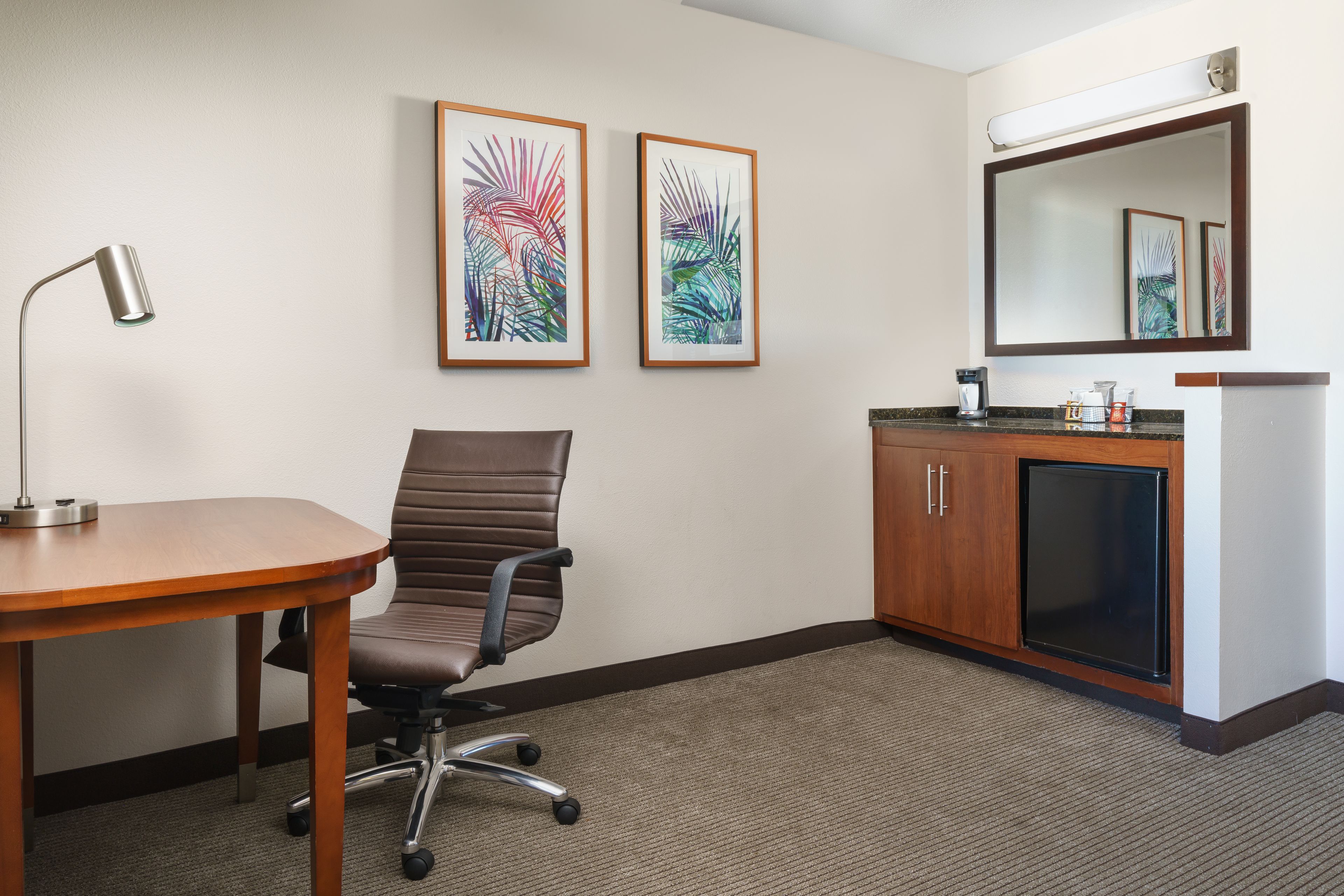 A hotel room with a wooden desk, a brown office chair, a flat-screen TV, a small cabinet, and two framed wall art pieces.