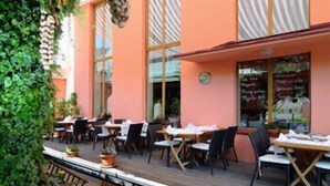 Ein Sitzbereich im Freien mit Tischen und Stühlen, eine Holzterrasse und ein Gebäude mit großen Fenstern und einer pfirsichfarbenen Fassade.