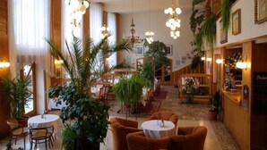 Eine großzügige Hotelhalle mit verschiedenen Sitzbereichen, darunter runde Tische und Sofas. Mehrere Kübelpflanzen und eine Kronleuchter sind an der Decke angebracht.