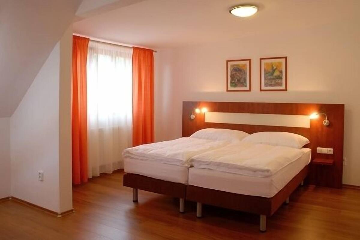 Une chambre avec un lit double, un plancher en bois, un plafond en pente et une fenêtre ornée de rideaux orange.