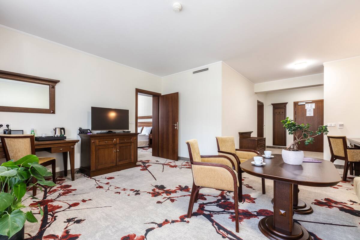 Ein Hotelzimmer mit Essbereich, Fernseher, Schreibtisch inklusive Computer und einer großen Topfpflanze.