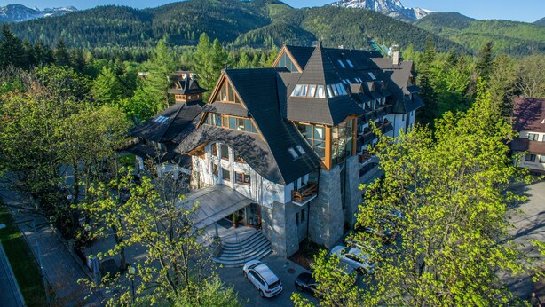 A large, multi-story house with a steep roof, surrounded by lush greenery and mountains in the background. - Hotel Crocus (Zakopane)