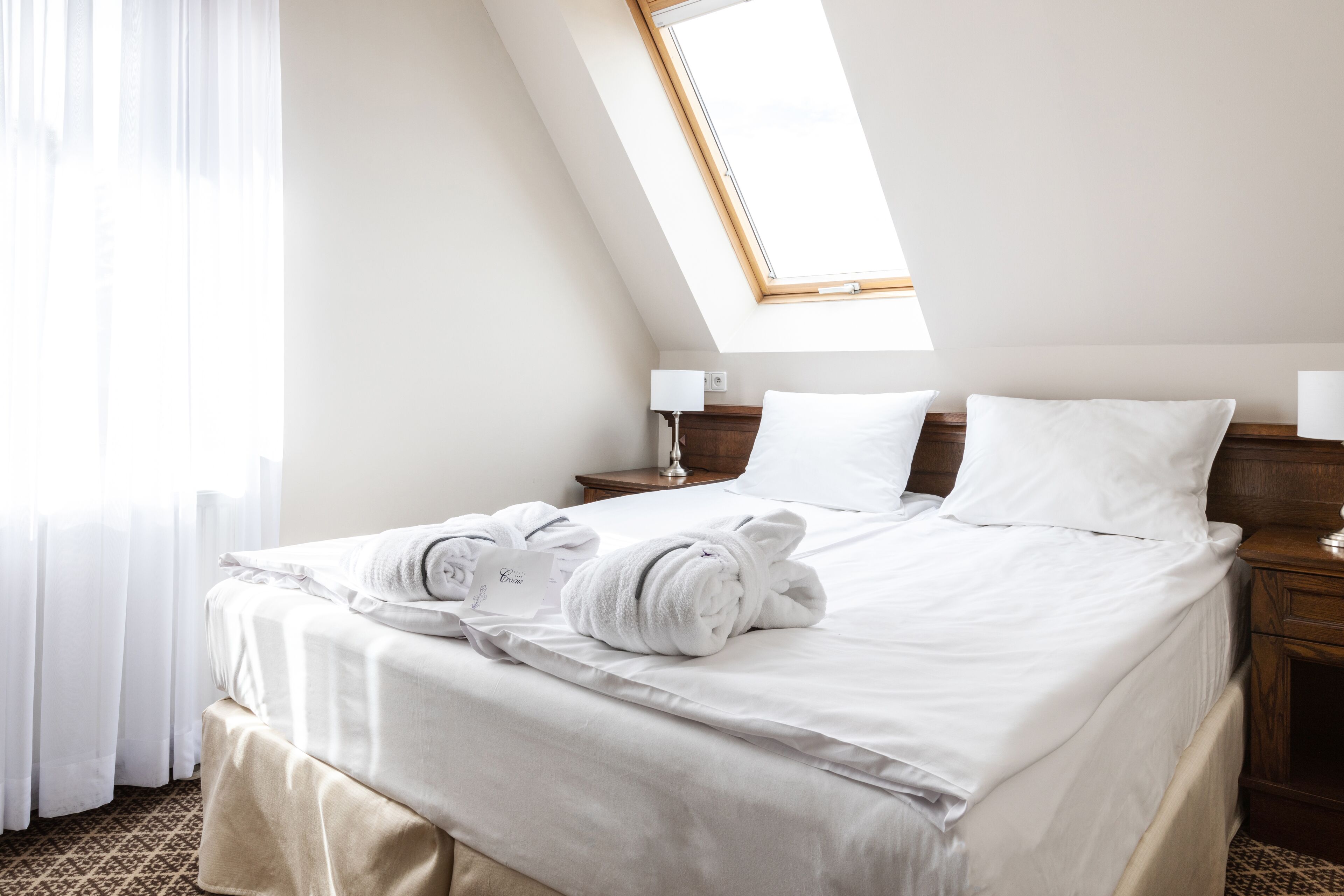 Una cama bien tendida con sábanas blancas y dos toallas, una mesita de noche con una lámpara, y una ventana con marco de madera.
