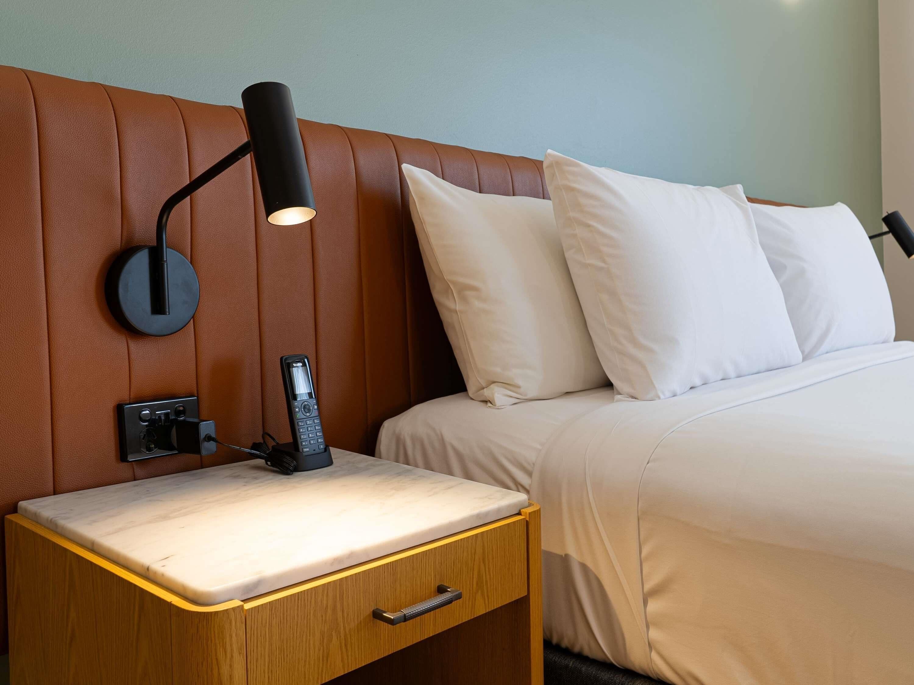 Une chambre d’hôtel avec un lit, une table de chevet avec une lampe, un téléphone et une petite table en bois.