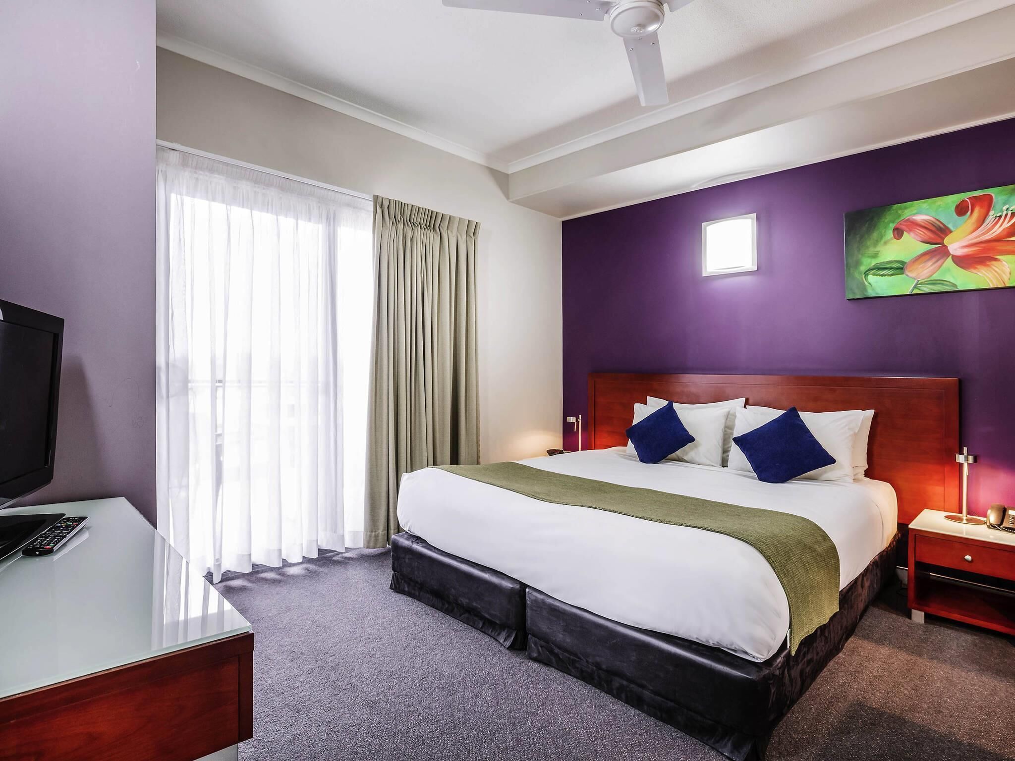 Una camera d'hotel con un letto, una scrivania con televisore, un comodino e una ventola a soffitto.
