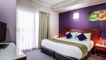Una camera d'hotel con un letto, una scrivania con televisore, un comodino e una ventola a soffitto.
