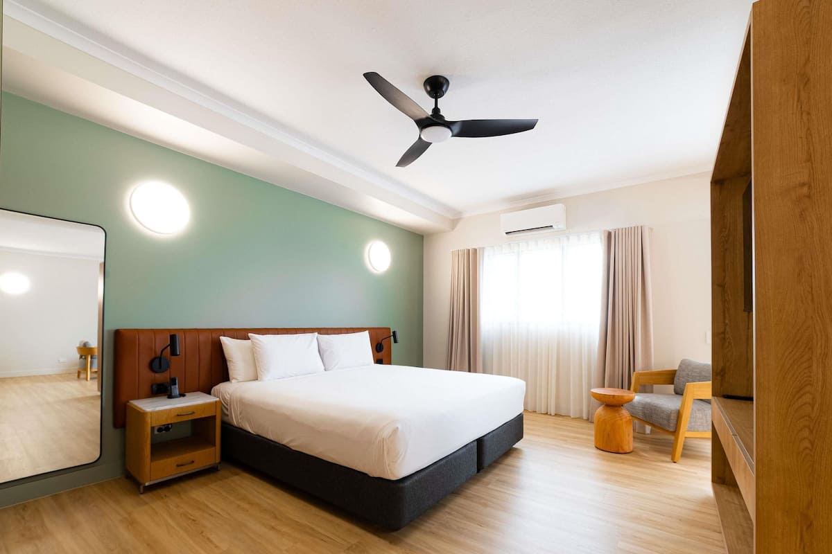 Habitación de hotel moderna con una cama grande, piso de madera, ventilador de techo, mesita de noche y vistas al exterior.