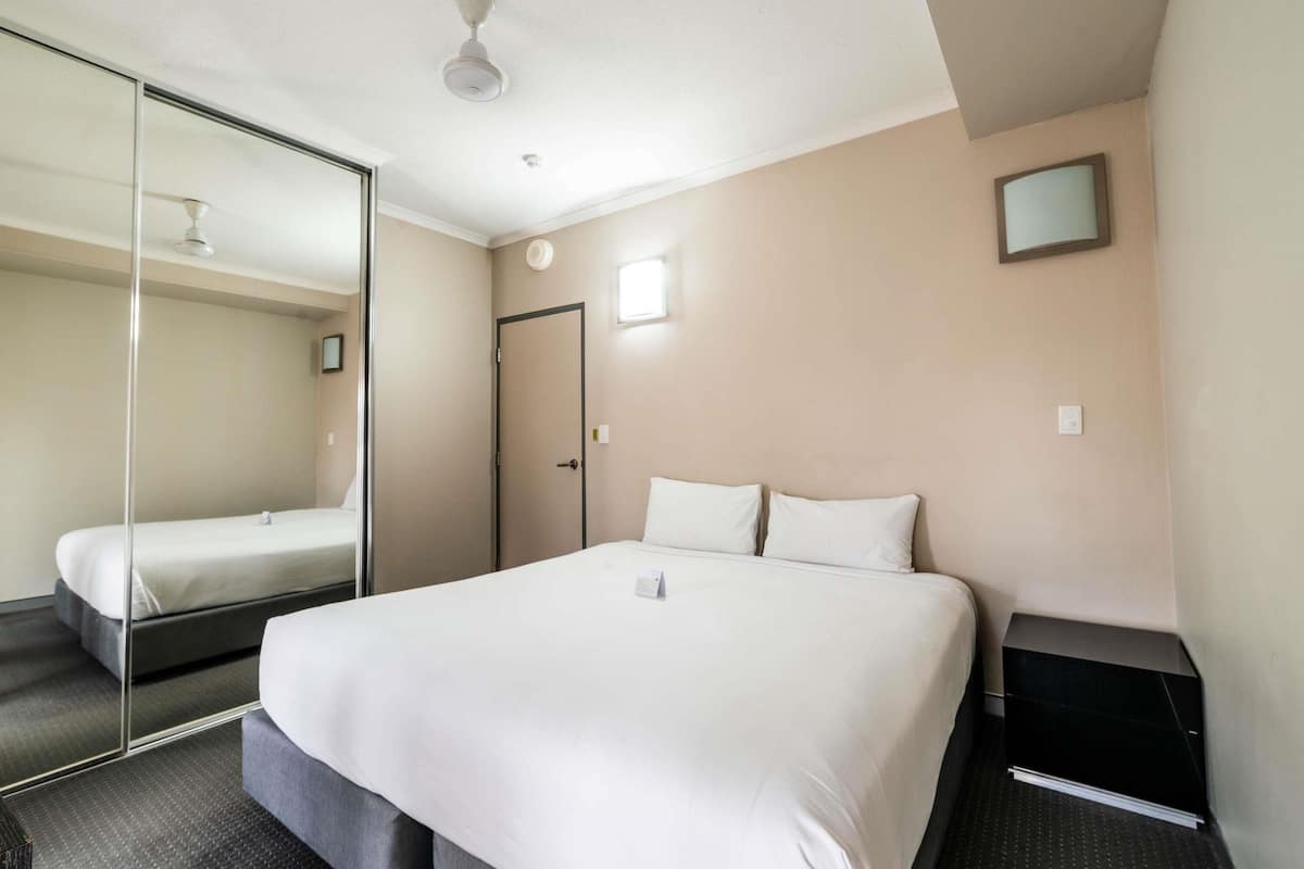 Una habitación de hotel con una cama, un espejo, un ventilador de techo, una puerta y una mesita de noche pequeña.