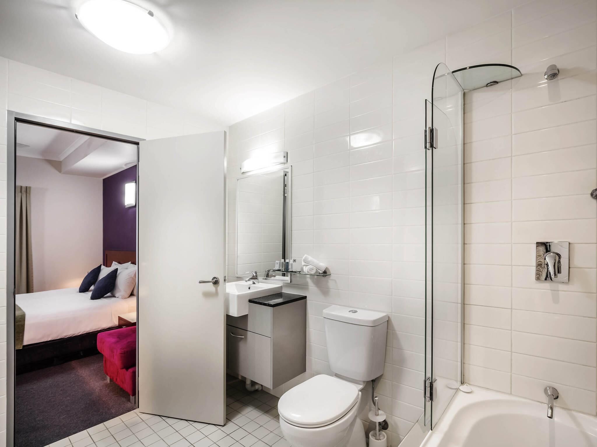 Uma casa de banho moderna com sanita, lavatório e chuveiro. Uma cama é visível no quarto adjacente.
