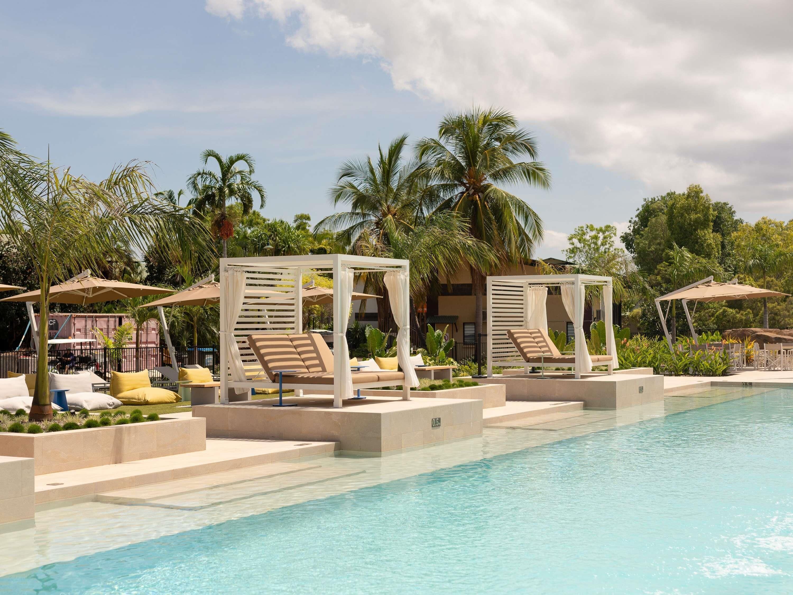 A poolside area with lounge chairs and umbrellas, surrounded by palm trees and a clear sky.