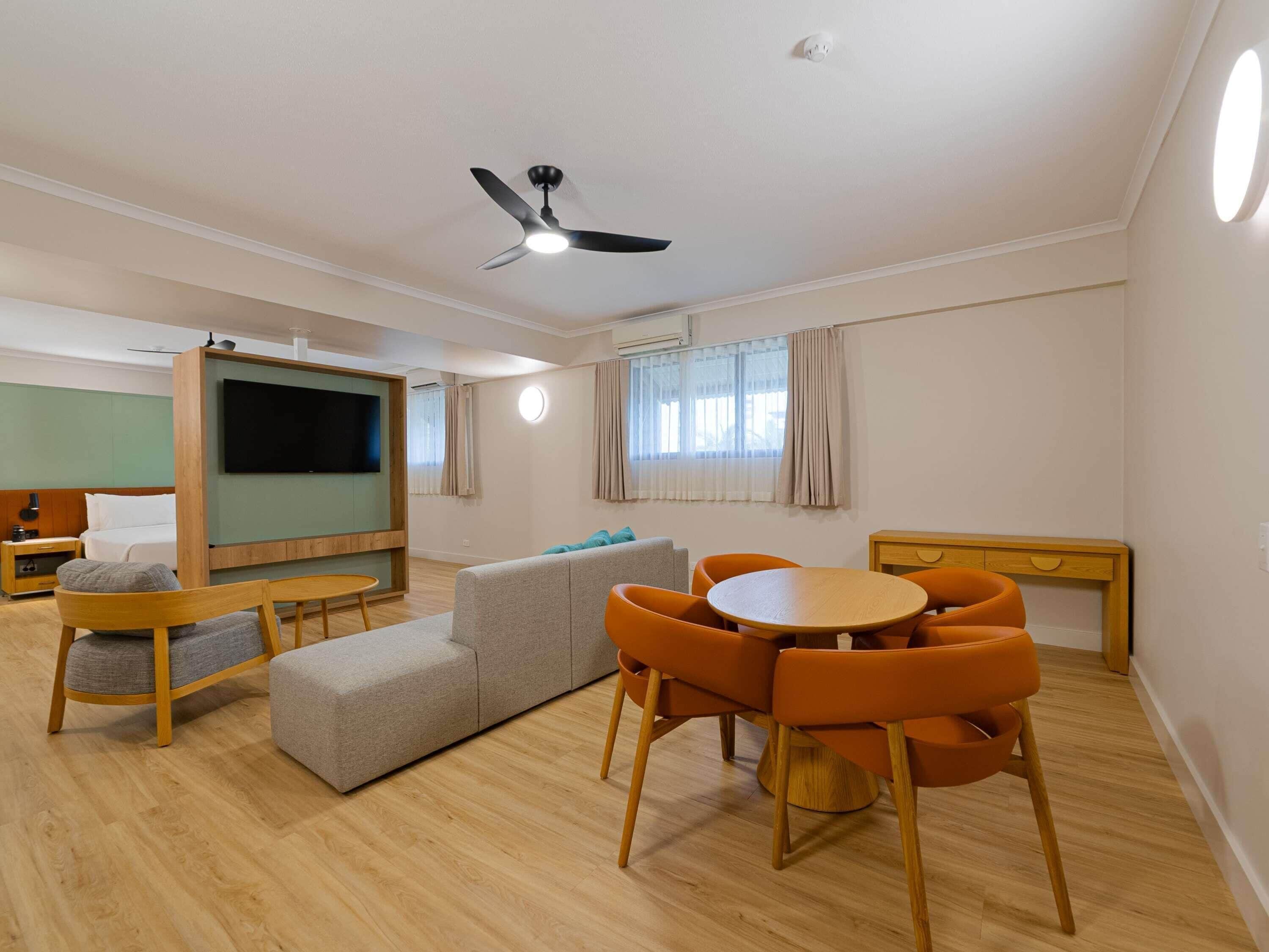 A modern hotel room with a flat-screen TV, a ceiling fan, a sofa, a dining table with chairs, and a small desk.