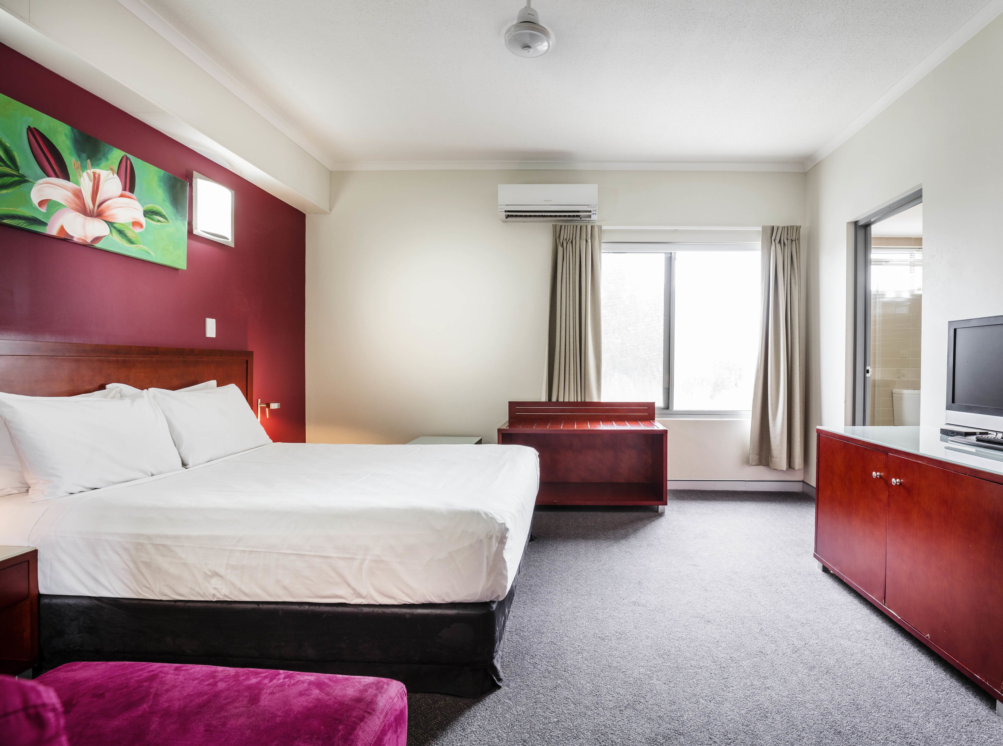 A hotel room with a bed, a desk, a TV, a window with curtains, and an air conditioning unit.