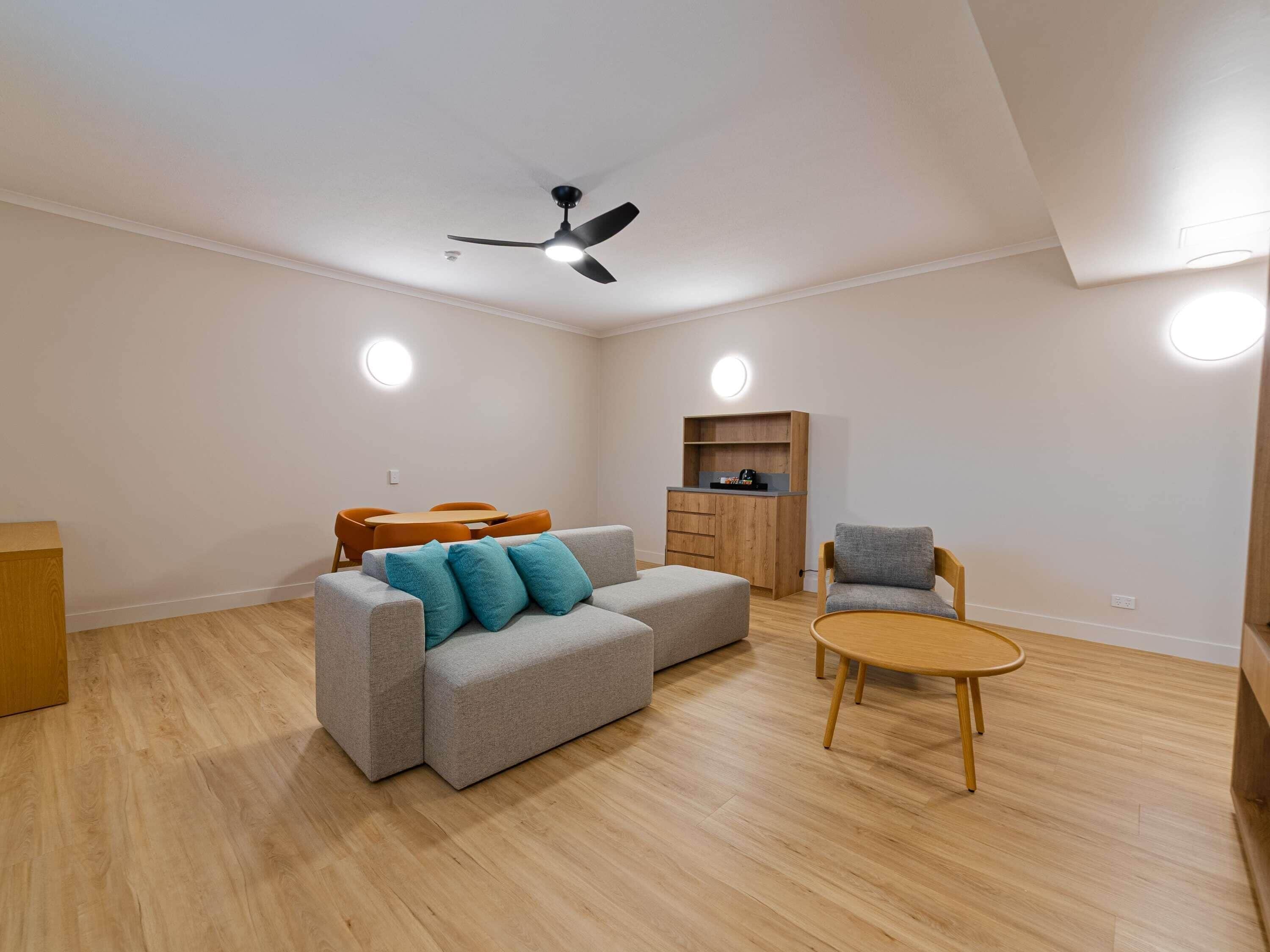 A modern living room with a grey sofa, a wooden coffee table, and a grey armchair. There is a wooden cabinet and a ceiling fan.
