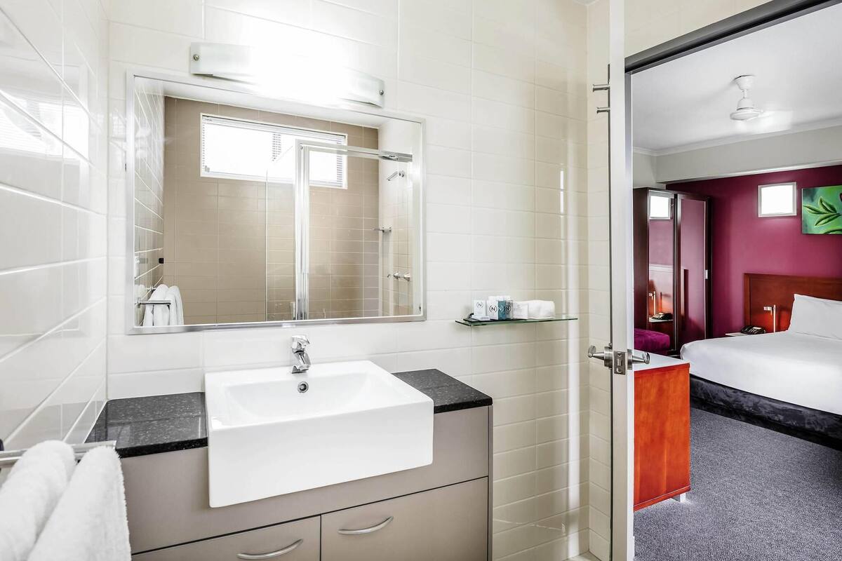 Baño de hotel con lavamanos blanco, espejo y cabina de ducha de vidrio.