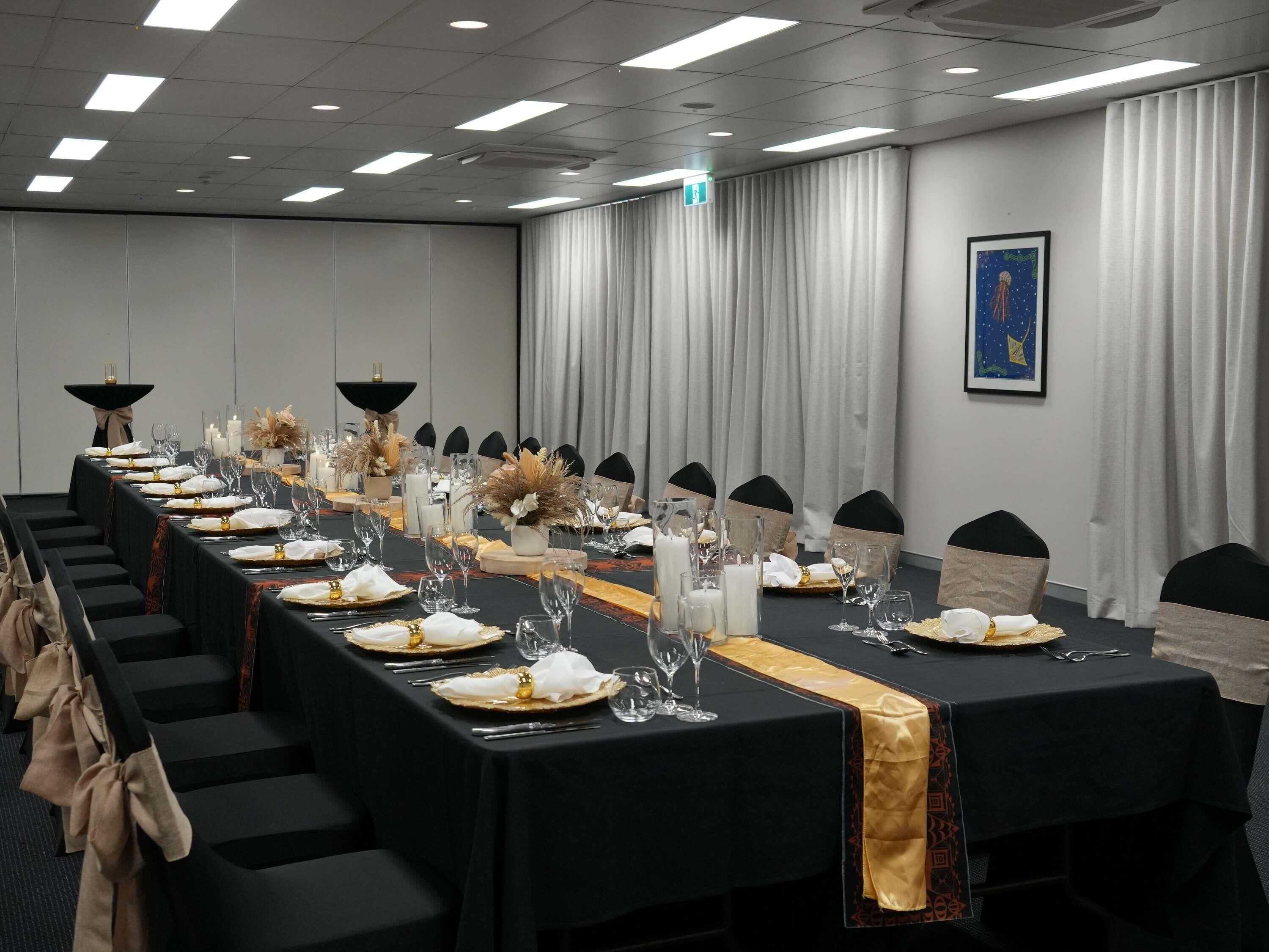 Un salón de conferencias con una larga mesa dispuesta para un evento formal, con sillas negras, manteles blancos y centros de mesa florales.