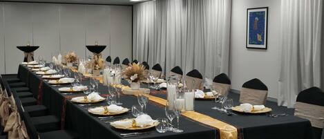 Un salón de conferencias con una larga mesa dispuesta para un evento formal, con sillas negras, manteles blancos y centros de mesa florales.