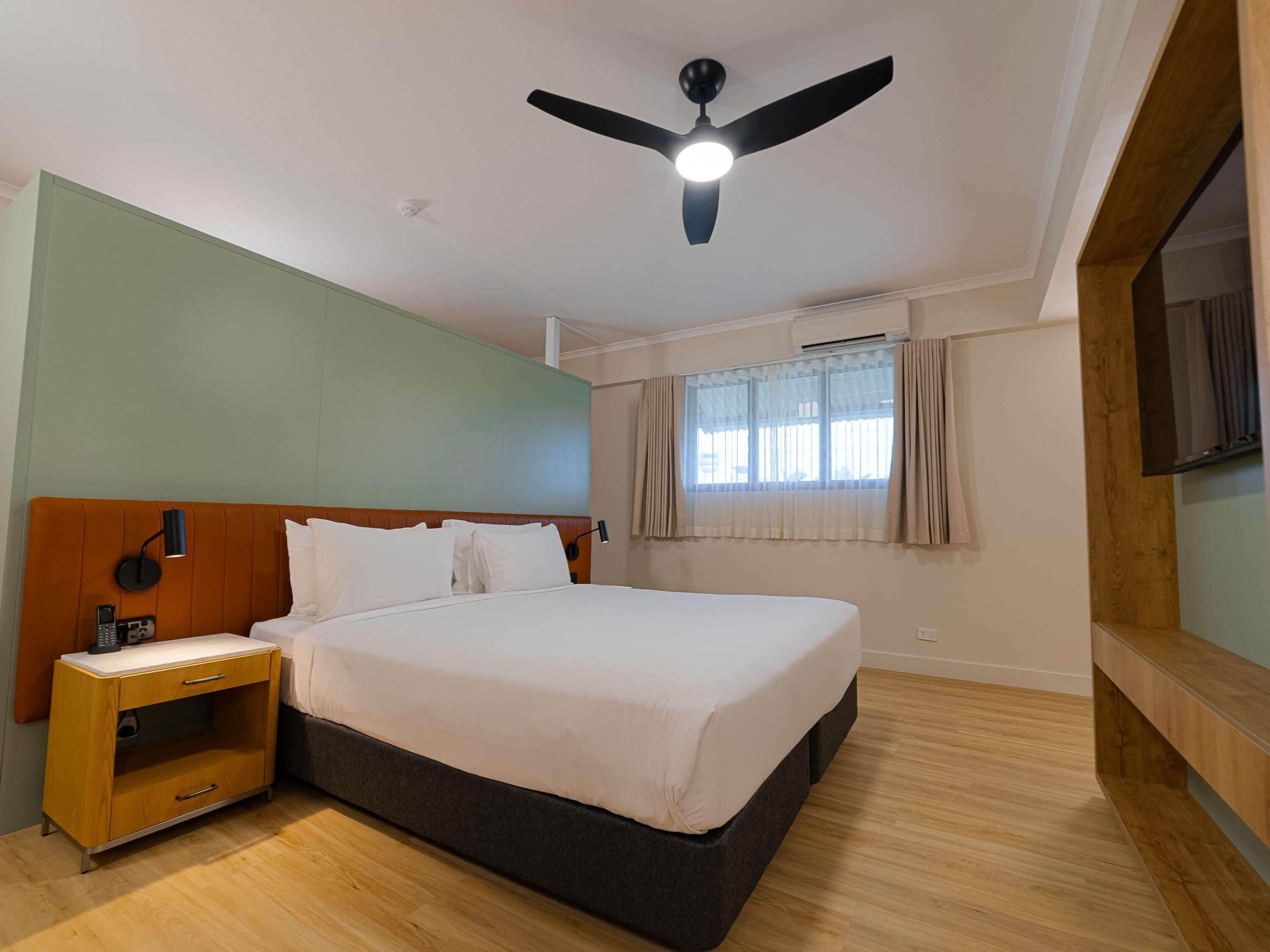 A hotel room with a bed, a nightstand, a ceiling fan, a window with curtains, and a TV mounted on the wall.