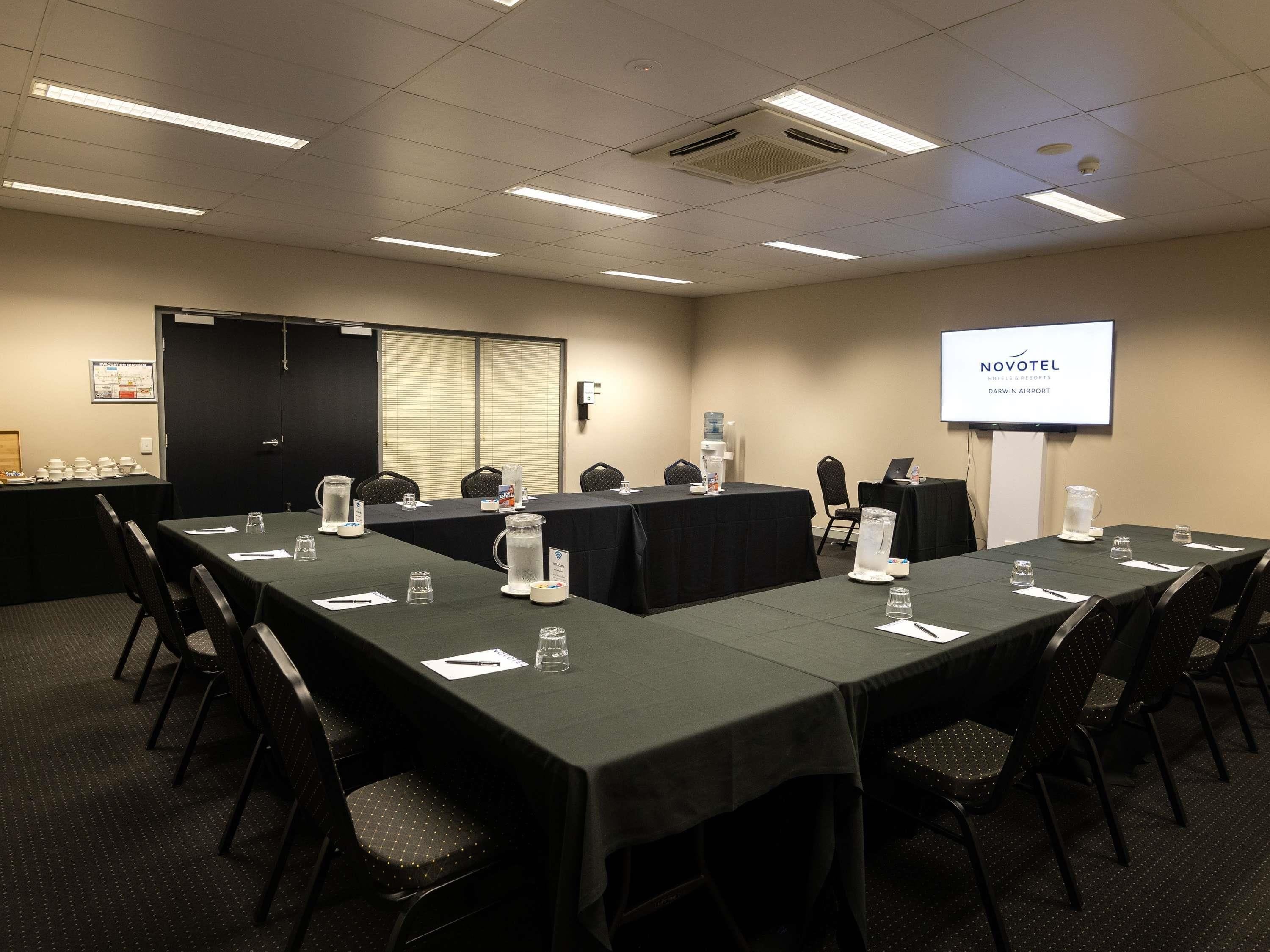 Une salle de conférence équipée de tables, de chaises et d’un écran de projection affichant le texte « NOVOTEL DARWIN AIRPORT ».