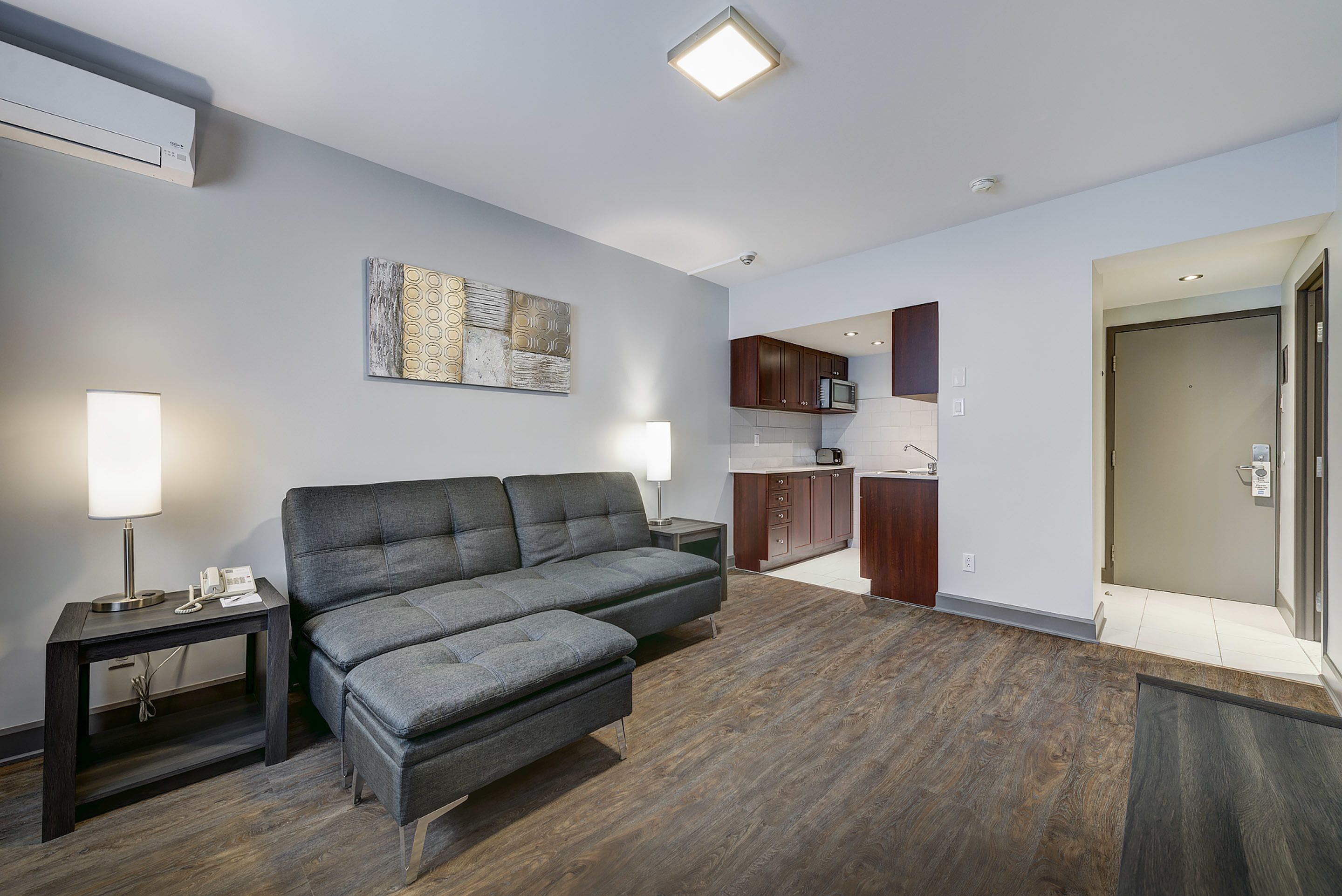 A modern living room with a grey sofa, a small table with a lamp, a kitchen area with dark cabinets, and a door leading to another room.