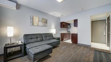 A modern living room with a grey sofa, a small table with a lamp, a kitchen area with dark cabinets, and a door leading to another room.