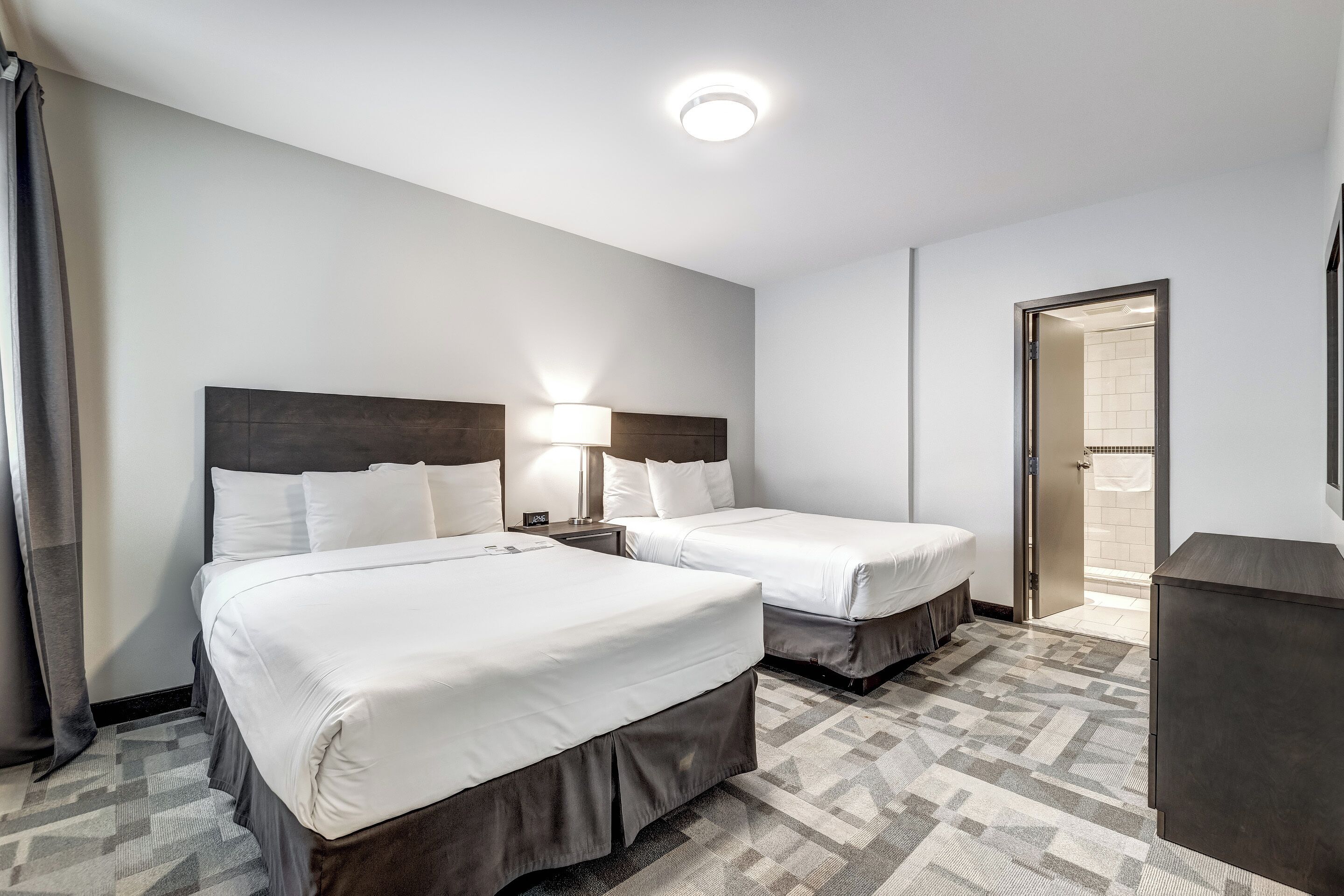 Habitación de hotel con dos camas, una mesita de noche, una lámpara, un aparador y la puerta del baño.