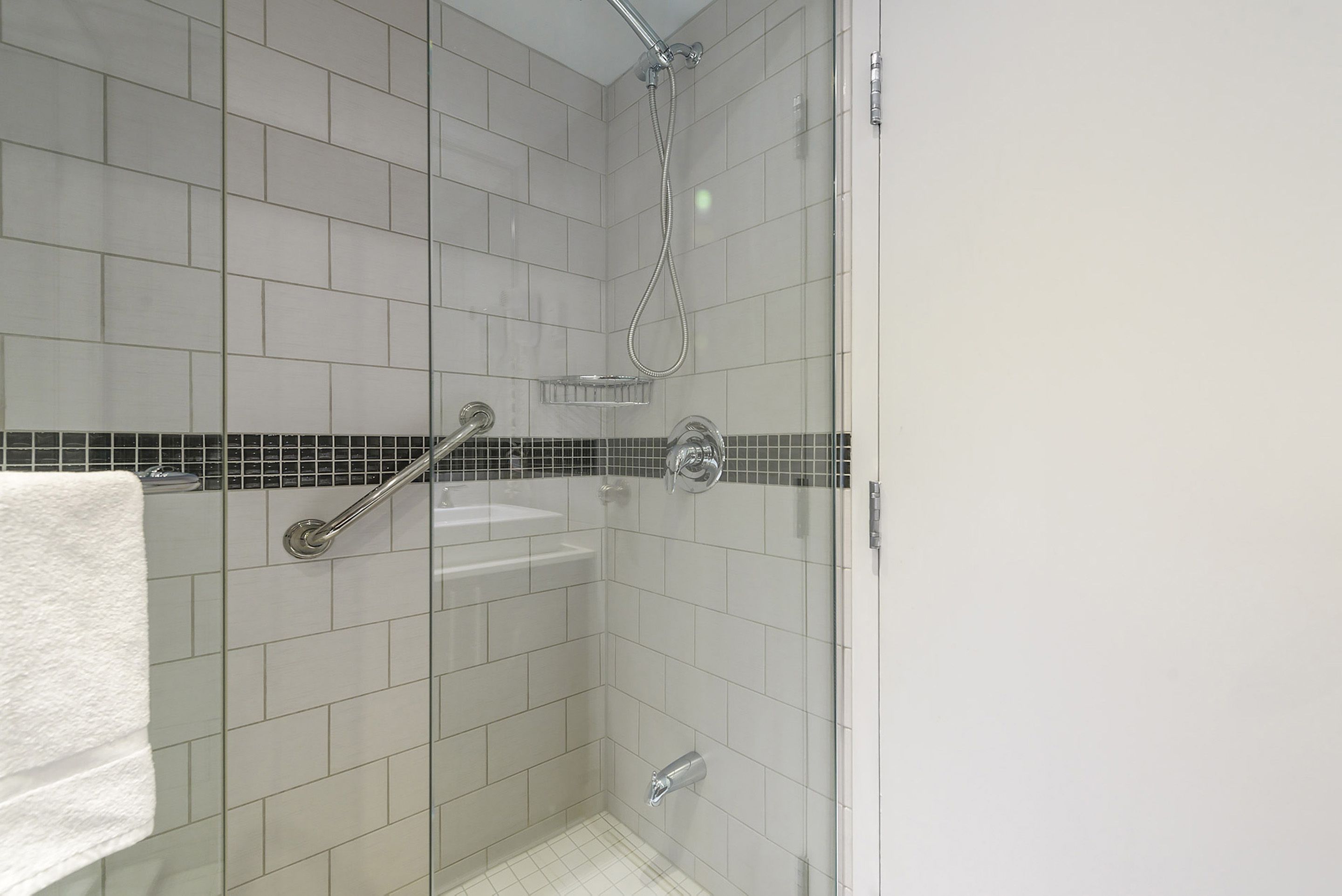 Une petite douche carrelée avec une porte en verre, une serviette blanche accrochée au mur et une barre d’appui en métal.