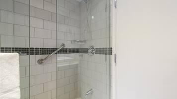 Une petite douche carrelée avec une porte en verre, une serviette blanche accrochée au mur et une barre d’appui en métal.