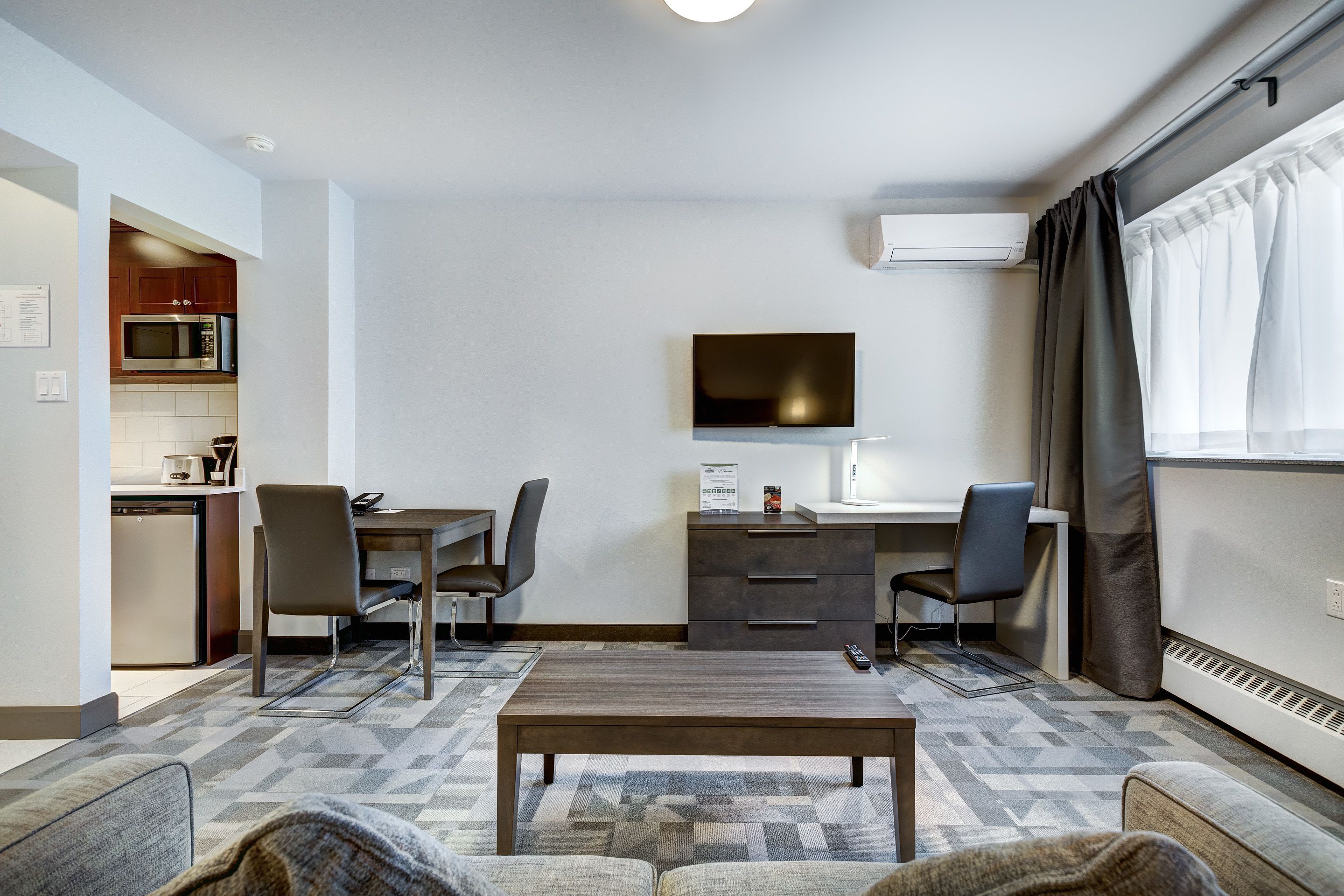 A compact hotel room with a kitchenette, a flat-screen TV, a desk with two chairs, and a sofa.