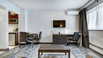 A compact hotel room with a kitchenette, a flat-screen TV, a desk with two chairs, and a sofa.