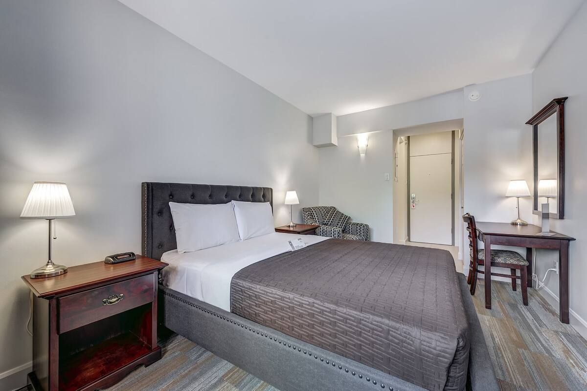 A hotel room with a large bed, a desk, a chair, and a nightstand with a lamp.