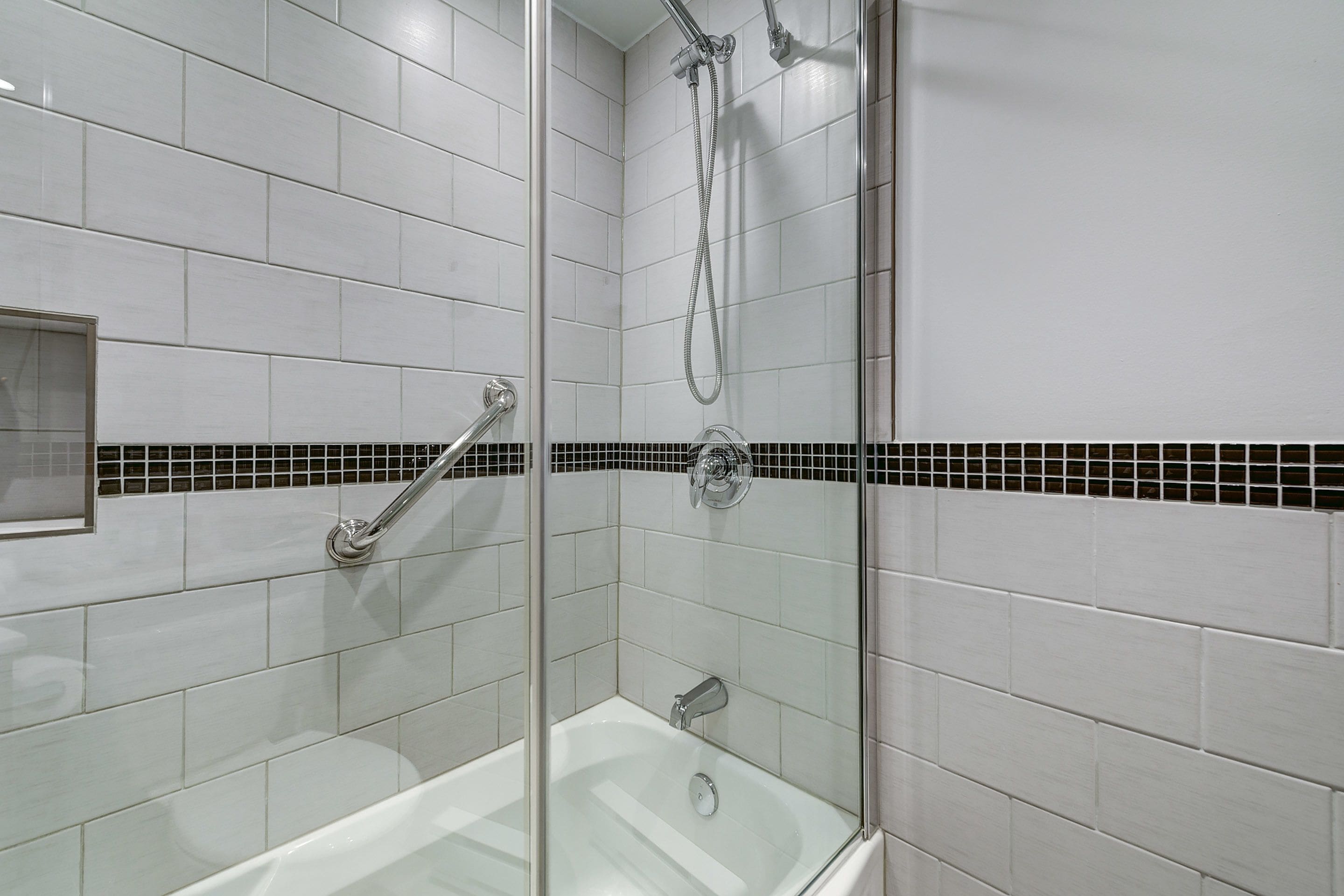Un baño moderno con azulejos blancos, ducha con cerramiento de vidrio y una franja decorativa de azulejos en blanco y negro.