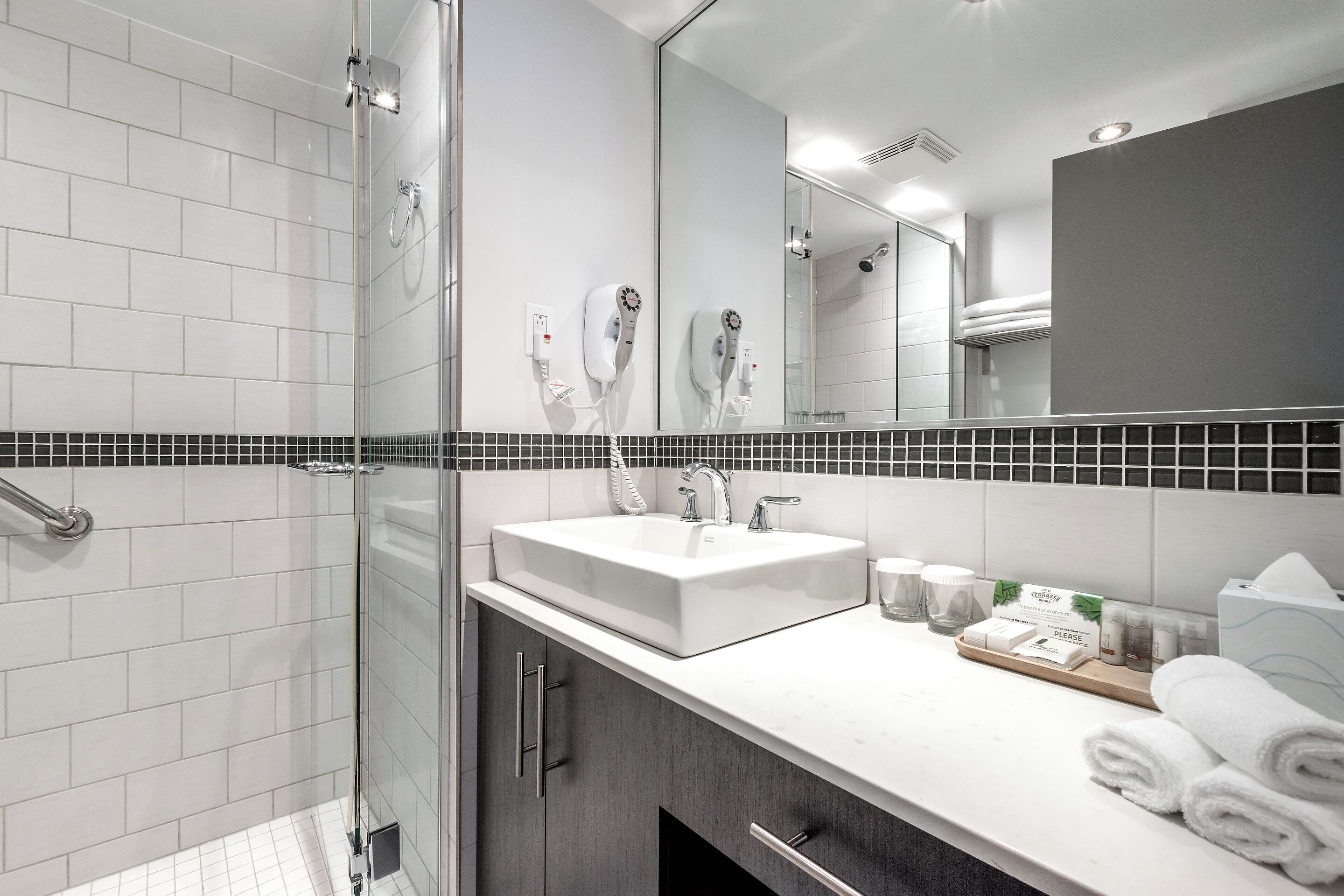 Un baño moderno con cabina de ducha de vidrio, un lavabo blanco con un espejo grande encima y un mueble de tocador con artículos de aseo.