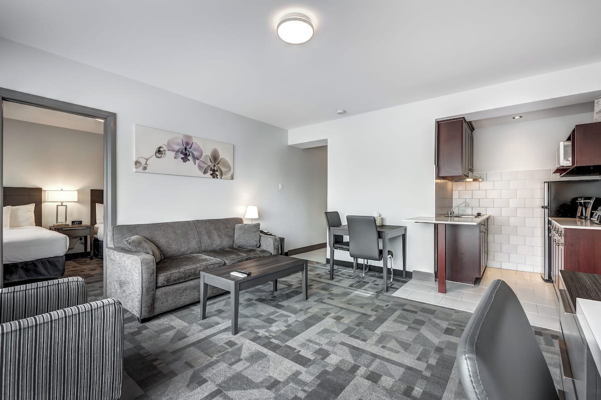 A modern hotel room with a sofa, armchair, dining table, and kitchen area.