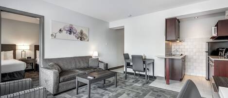 A modern hotel room with a sofa, armchair, dining table, and kitchen area.
