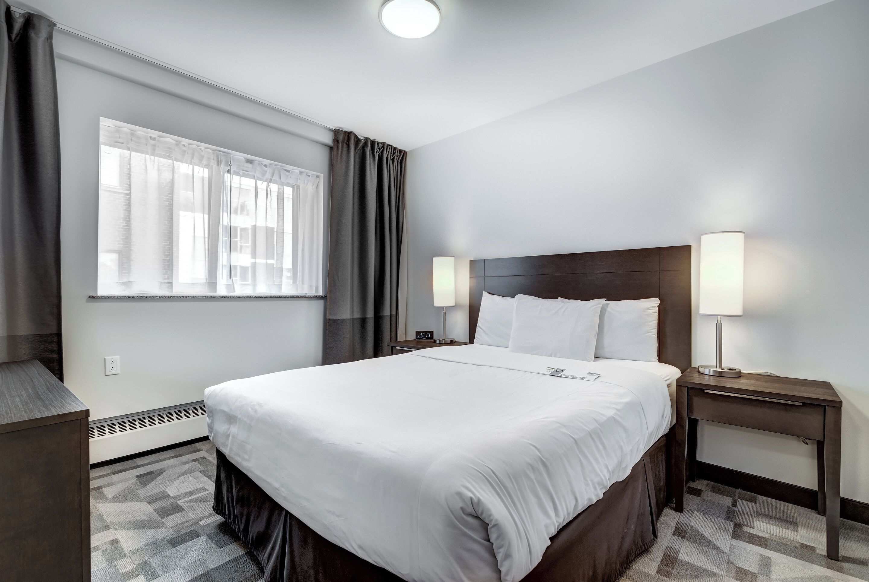 Ein Hotelzimmer mit einem großen Bett, zwei Nachttischen mit Lampen, einem Fenster mit Vorhängen und einem Heizkörper.