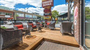 An outdoor seating area with wicker furniture and red cushions, a digital sign displaying room rates, and a view of the street with various shops. - Hotel Terrasse Royale (Montreal)