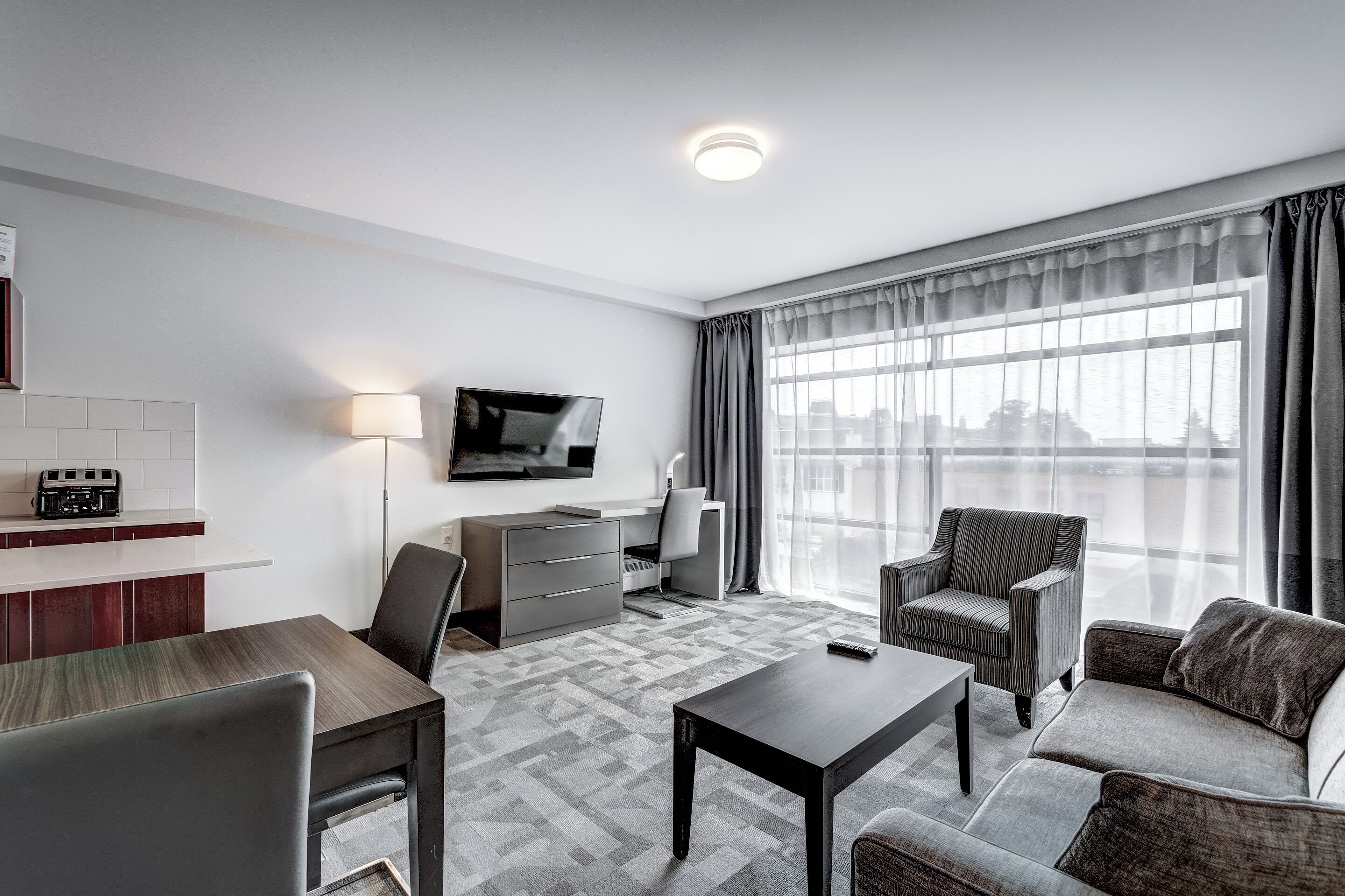 A modern hotel room with a dining area, a sofa, a TV, and a large window with curtains.