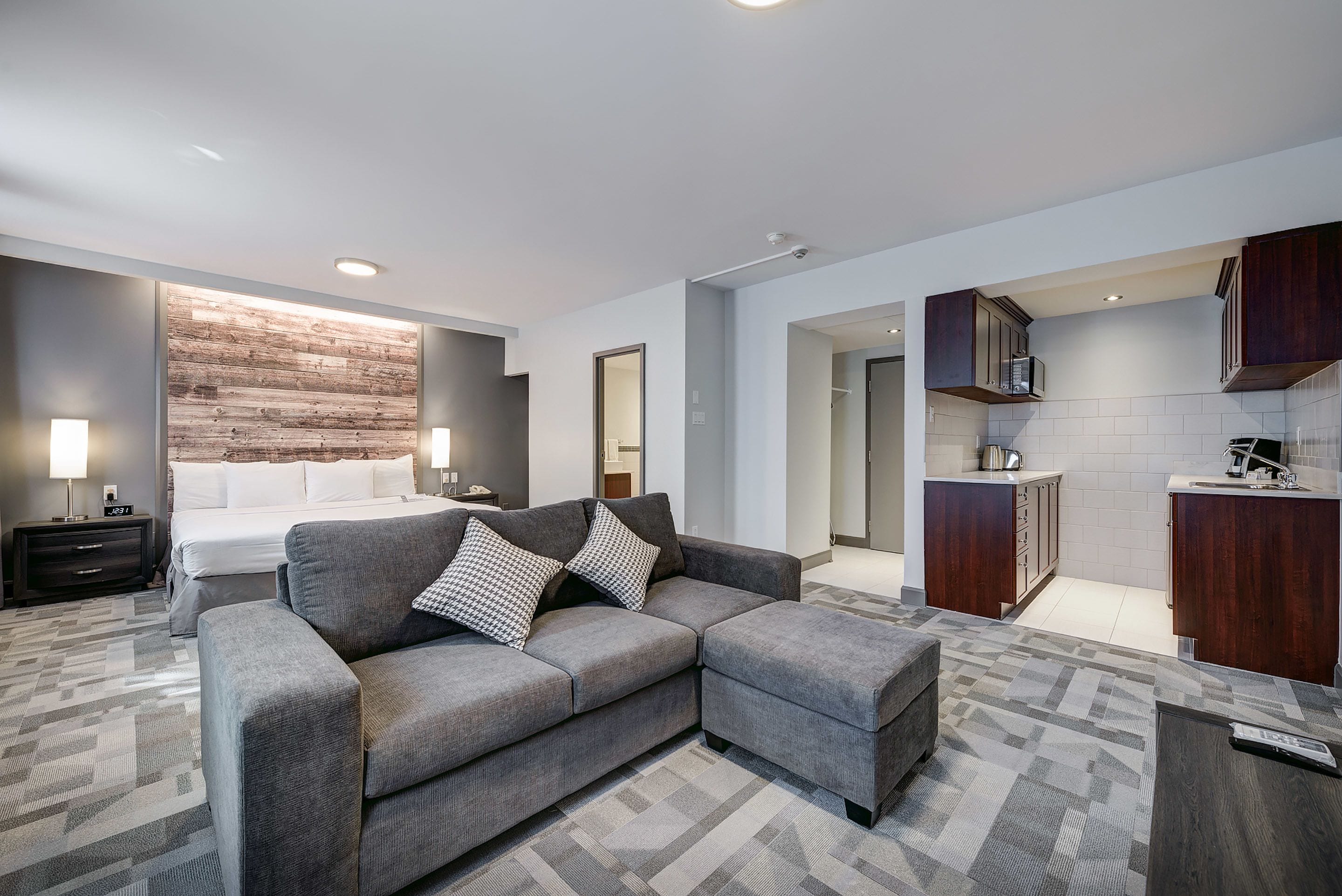 Une chambre d’hôtel moderne avec un canapé d’angle gris, un lit avec du linge de maison blanc, un mur d’accent en bois, une kitchenette avec des meubles en bois foncé et un tapis à motifs.